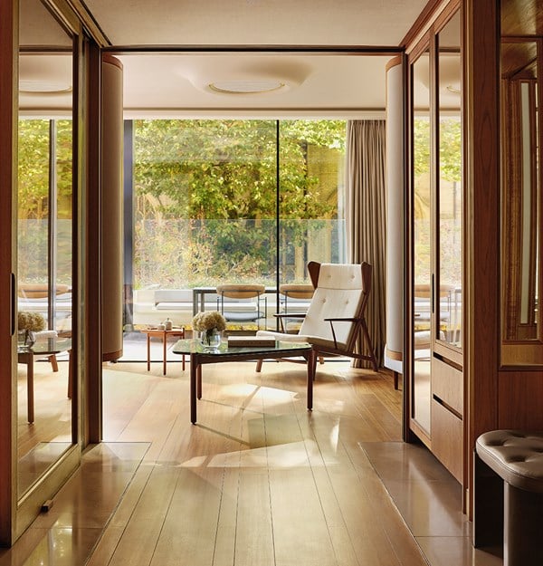 View from the entrance of a bright and elegant hotel suite featuring wood flooring, a modern armchair with white upholstery, a glass coffee table with flowers and books, and floor-to-ceiling windows opening onto a private terrace with greenery and outdoor seating.