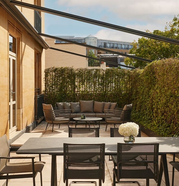 Spacious terrace featuring a modern dining table with mesh chairs and a separate seating area with cushioned armchairs and a sofa, surrounded by tall greenery and partial views of urban buildings.