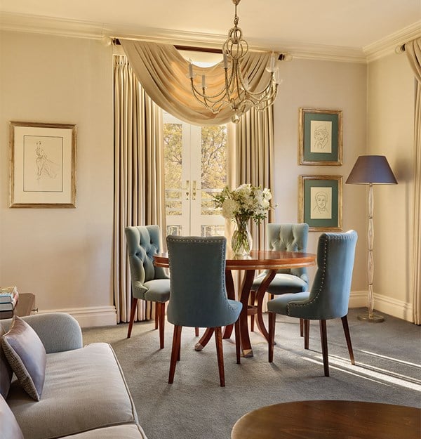 Refined dining area with a round wooden table and blue velvet chairs beneath a chandelier, set against cream walls with framed art and tall draped curtains framing French doors to a balcony.