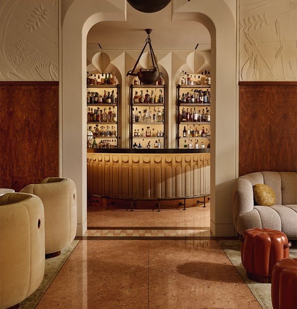 A warmly lit bar with an arched design, upholstered armchairs, and shelves filled with bottles behind a curved counter.
