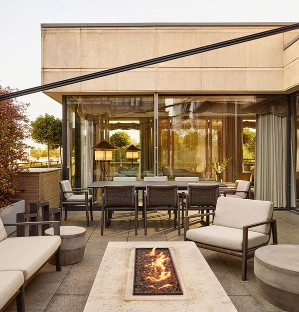 A spacious terrace with lounge seating, dining table, and a central fire feature, framed by glass walls and greenery.