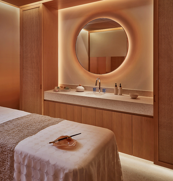 A softly lit spa treatment room with a massage table, warm wooden cabinetry, and a large round mirror above a sink.