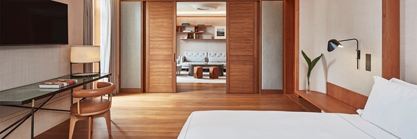 A modern, minimalist suite with a large bed, a wall-mounted TV, a sleek desk with a chair, and a view into a cosy sitting area with a sofa and bookshelves through sliding wooden doors. The room features wooden floors and neutral-toned decor.