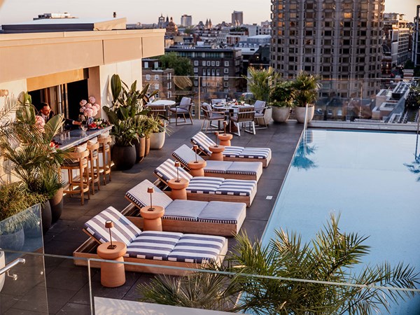 A rooftop pool area features striped lounge chairs, potted plants, and a bar with stools. In the background, there are dining tables set with meals, creating a relaxing and inviting atmosphere.