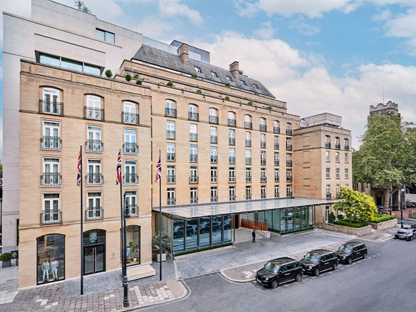 The exterior of The Berkeley with beige stone walls, multiple flags flying outside, and several black taxis parked in front. The entrance is covered by a glass canopy, and there is a doorman standing at the steps leading inside.