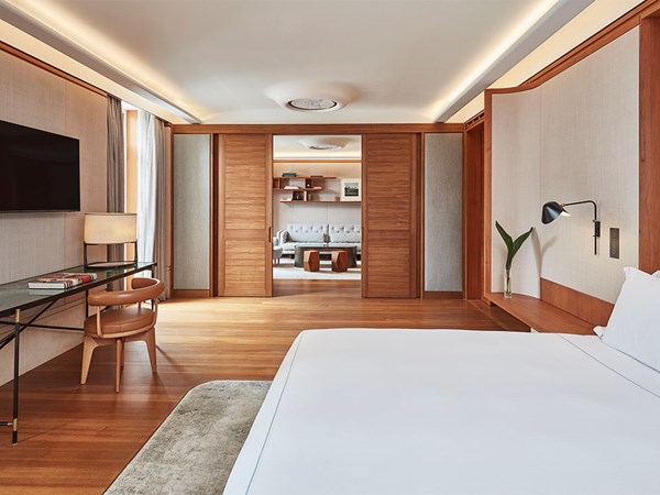 A modern, minimalist suite with a large bed, a wall-mounted TV, a sleek desk with a chair, and a view into a cosy sitting area with a sofa and bookshelves through sliding wooden doors. The room features wooden floors and neutral-toned decor.