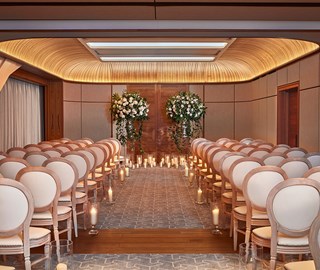 An aisle in an indoor event space laid out for a wedding with row of chairs, candles and flowers.