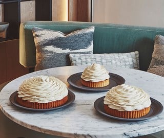 Three round cakes topped with swirls of white cream are displayed on black plates on a marble table in a modern lounge setting.