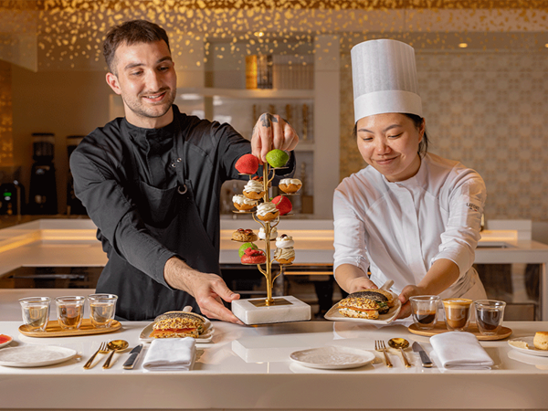 Two pastry chefs smiling as they present an intricate golden tree stand filled with colourful pastries and serve gourmet sandwiches and coffee at a pristine white chef’s counter under warm lighting.