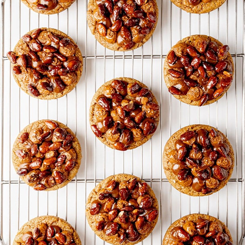 Twelve round praline cookies topped with glossy caramelised nuts, evenly spaced on a wire cooling rack against a bright white surface.