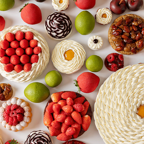 Overhead view of artistic pastries with whipped cream, berries, and citrus desserts arranged alongside limes and trompe-l’œil fruit on a white surface.