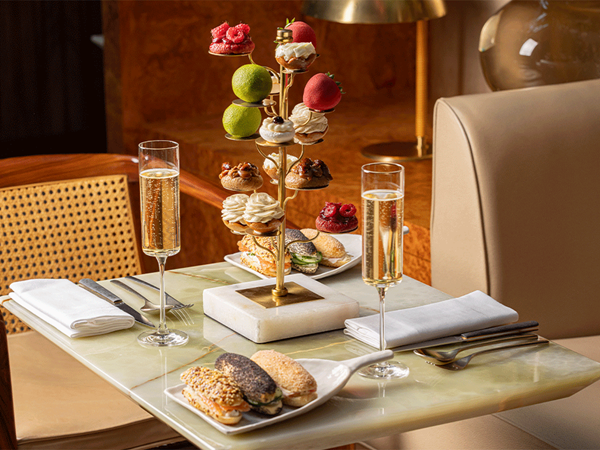 Elegant Four Goutea tea service featuring a marble table set with sandwiches, champagne, and a sculptural gold stand showcasing fine Cedric Grolet pastries.