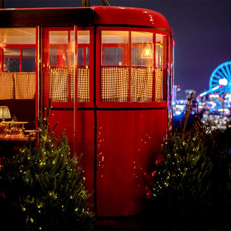 A charming red cable car cabin illuminated from within, surrounded by twinkling fairy lights and miniature Christmas trees, with a vibrant Ferris wheel glowing in the background.