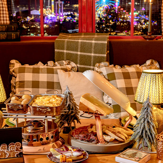 An elegant alpine-inspired dining setup featuring a raclette station, copper serving dishes, and a cozy arrangement of plaid cushions and mini pine tree decorations, with a vibrant cityscape visible through the window