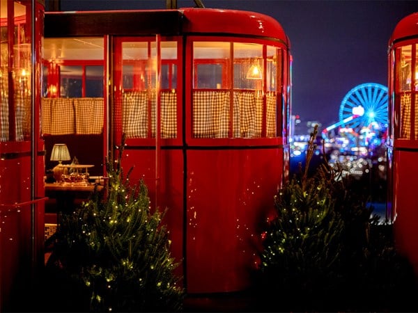 Red ski gondola-style dining cabins with festive lights, overlooking a city skyline and Ferris wheel at night.
