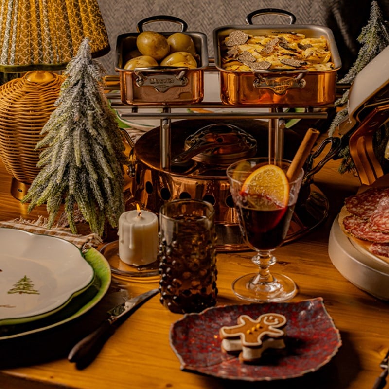 A cozy holiday dining setup featuring copper serving dishes, mulled wine garnished with orange and cinnamon, a gingerbread cookie, and a festive, rustic table adorned with miniature snow-covered trees and warm candlelight.