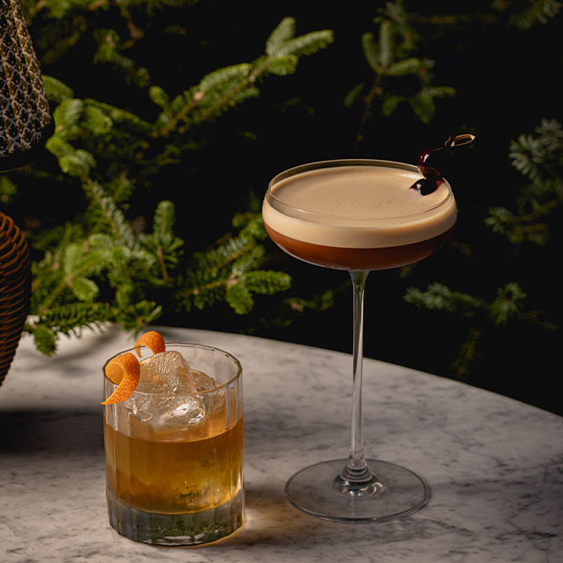 An elegant cocktail pairing of a Negroni and an Espresso Martini, served against a backdrop of evergreen branches, offering a refined and festive ambiance for a winter celebration.