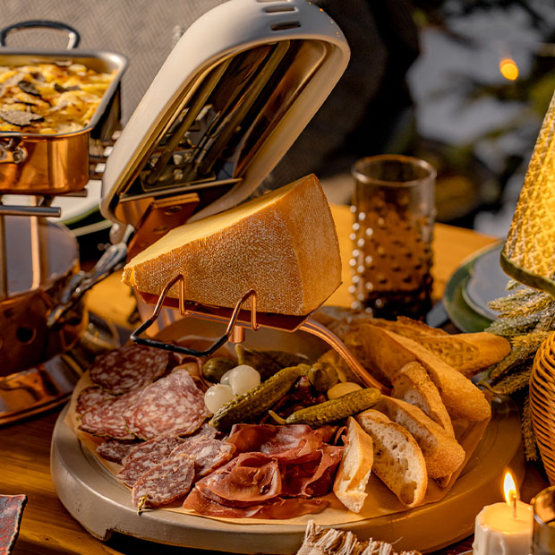 A cozy raclette station featuring a wheel of cheese melting alongside a beautifully arranged charcuterie board with cured meats, pickles, and slices of rustic bread, setting the stage for an indulgent Alpine feast.