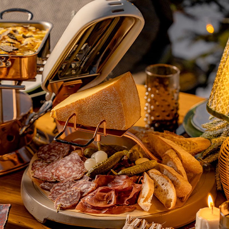 A cozy raclette station featuring a wheel of cheese melting alongside a beautifully arranged charcuterie board with cured meats, pickles, and slices of rustic bread, setting the stage for an indulgent Alpine feast.