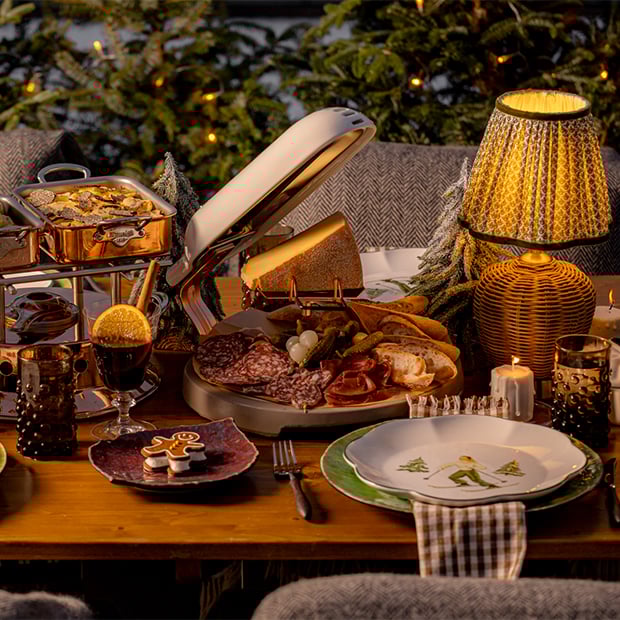 An intimate and festive dining scene featuring a raclette cheese wheel, charcuterie board, copper serving dishes, and mulled wine, set against a backdrop of twinkling lights and snow-dusted evergreens, creating a warm, alpine-inspired atmosphere.