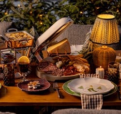 An intimate and festive dining scene featuring a raclette cheese wheel, charcuterie board, copper serving dishes, and mulled wine, set against a backdrop of twinkling lights and snow-dusted evergreens, creating a warm, alpine-inspired atmosphere.