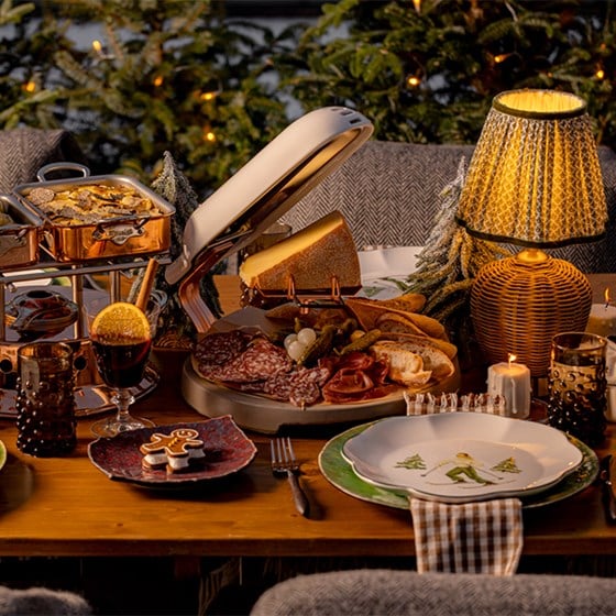 An intimate and festive dining scene featuring a raclette cheese wheel, charcuterie board, copper serving dishes, and mulled wine, set against a backdrop of twinkling lights and snow-dusted evergreens, creating a warm, alpine-inspired atmosphere.
