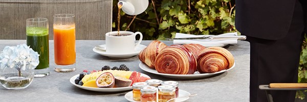Outdoor breakfast table with croissants, tropical fruit, juice, tea being poured, and mini jams beside a blue hydrangea flower.