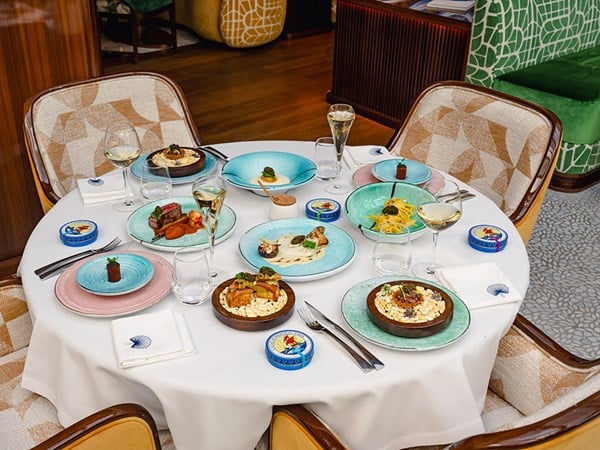 A round white table set with an eight-course caviar tasting menu, featuring colourful plates of gourmet dishes, Petrossian caviar tins, and glasses of white wine and champagne, in a stylish restaurant setting.