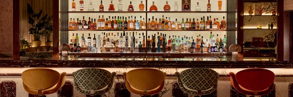 Elegant hotel bar with marble counter, five velvet chairs, and a backlit display of premium spirits in glass shelves.