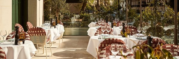 Sunny terrace of La Môme restaurant with white tablecloths, glassware, and red-checkered chairs surrounded by greenery and glass railing.