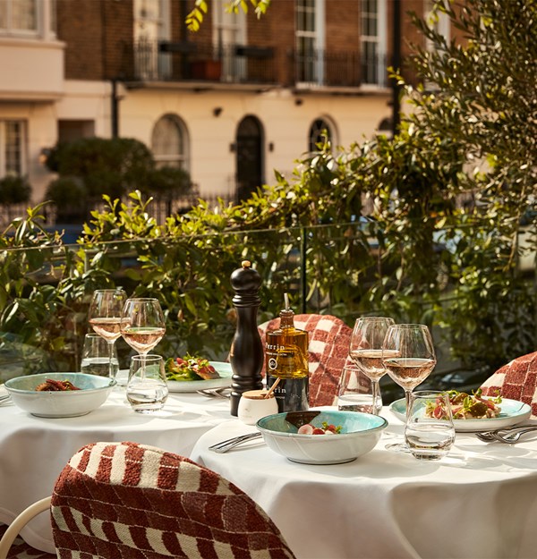 Close-up of a sunlit restaurant terrace table set with wine glasses, salads, and condiments, surrounded by red-checkered chairs and leafy greenery.