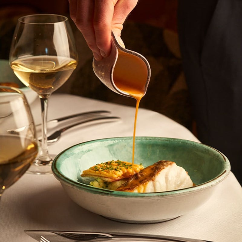 A delicate pour of golden sauce over a beautifully plated dish of white fish and crispy garnish, paired with a glass of white wine for a refined dining experience.