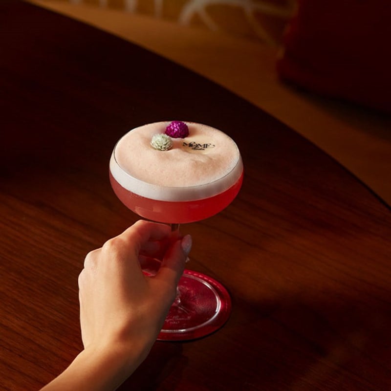 A hand gracefully holding a vibrant cocktail with a frothy top, adorned with delicate edible flowers and bespoke branding, set against a polished wooden table.