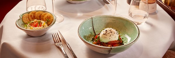 Burrata dish on a white tablecloth, served with diced vegetables and a side bowl of dip with small round crackers.