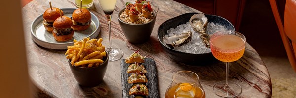 A marble table at The Berkeley Bar set with wagyu sliders, truffle fries, oysters on ice, yellowtail cones, mini lobster tacos, and five signature cocktails.