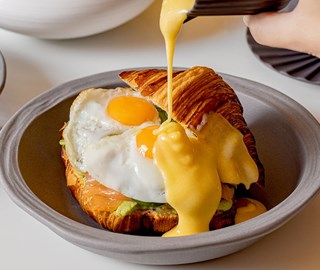 Hollandaise sauce being poured into croissant stuffed with eggs, smoked salmon and avocado