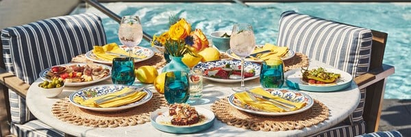 Poolside dining setup with Italian dishes, turquoise glasses, and striped cushions.
