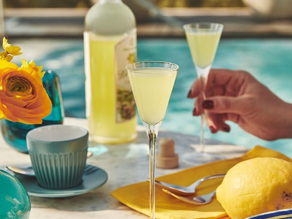 Hand holding a limoncello glass beside a pool with lemons and tableware nearby.
