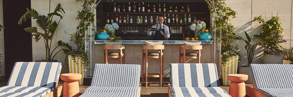 Bartender behind a rooftop bar with colorful drinks, striped loungers in foreground, and vibrant plants decorating the scene.