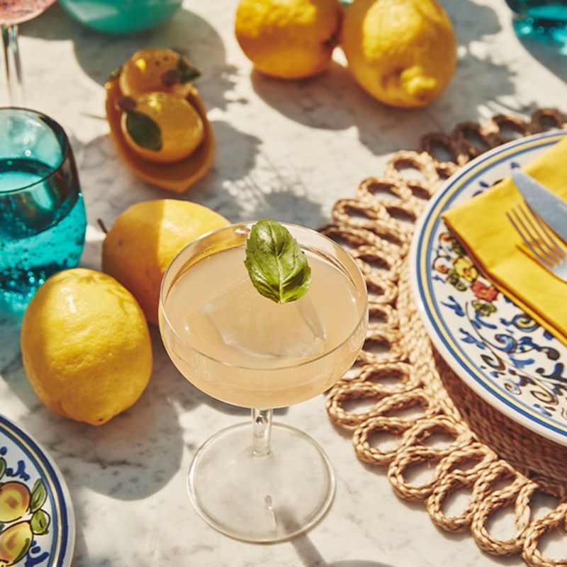 Citrus cocktail with ice and a basil leaf, surrounded by lemons and vibrant tableware.