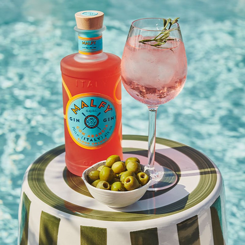 Malfy gin bottle, pink cocktail with rosemary, and green olives by a poolside.