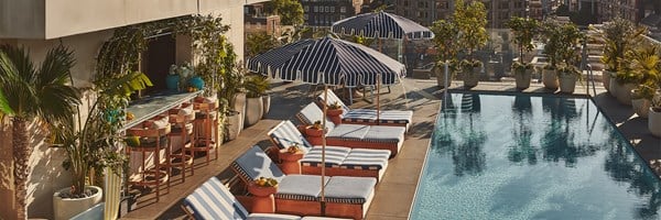 Rooftop pool area with blue striped loungers, umbrellas, a marble bar with stools, lush potted plants, and cityscape views under clear blue skies.