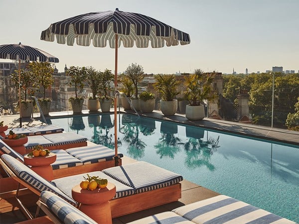 Sunlit rooftop pool with striped loungers, umbrellas, citrus fruit bowls, and surrounding potted plants, overlooking a green park and distant city skyline.