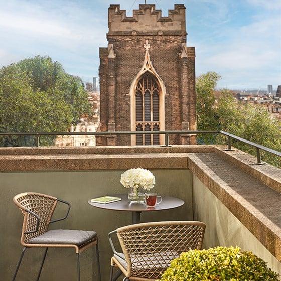 Private balcony of the Balcony Junior Suite at The Berkeley, featuring a round table with two woven chairs, a vase of white flowers, and a view of a historic church tower framed by trees and the London skyline