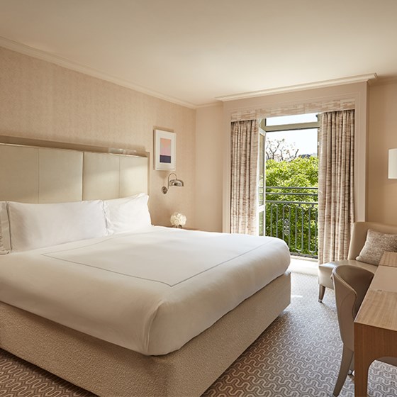 Bright hotel bedroom with a king-sized bed, neutral tones, a writing desk, and French doors opening to a Juliet balcony with garden views.