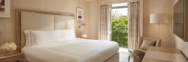 Bright hotel bedroom with a king-sized bed, neutral tones, a writing desk, and French doors opening to a Juliet balcony with garden views.