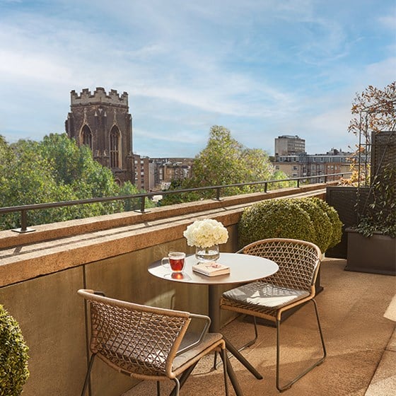 Sunny private balcony with a city view, featuring a round table with two chairs, flowers, and a drink.