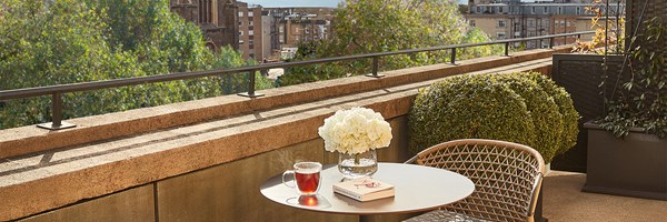 Balcony with a round table, two woven chairs, a cup of tea, a book, and a vase of white flowers overlooking city rooftops and trees.
