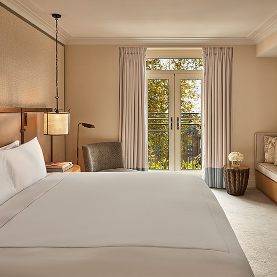 Contemporary hotel room with a king-sized bed, soft neutral tones, a lounge chair, and French doors opening to a Juliet balcony.