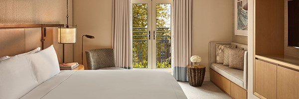 Contemporary hotel room with a king-sized bed, soft neutral tones, a lounge chair, and French doors opening to a Juliet balcony.
